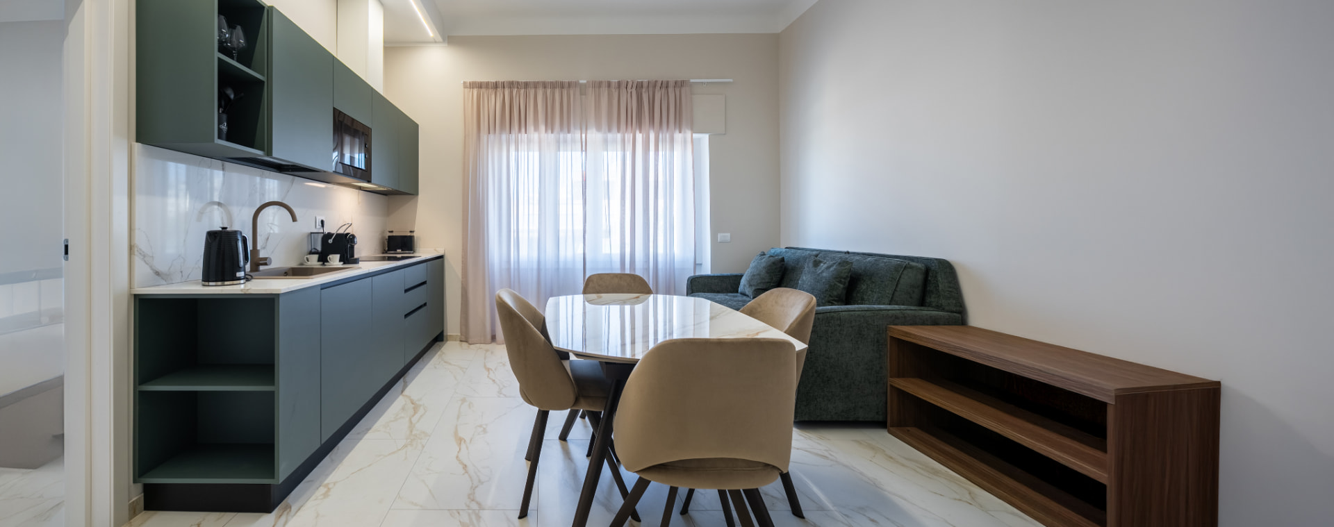 Appartamento luminoso con cucina a vista verde salvia, tavolo da pranzo con poltroncine beige e zona living con finestra pannellata da tende leggere
